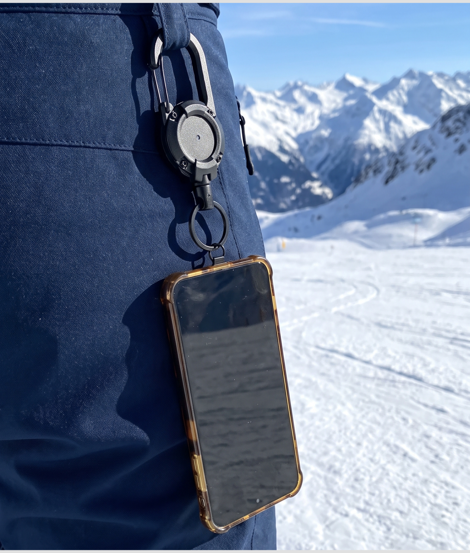 Telefoon aan een koord met karabijnhaak aan een skibroek, bergen en sneeuw op de achtergrond
